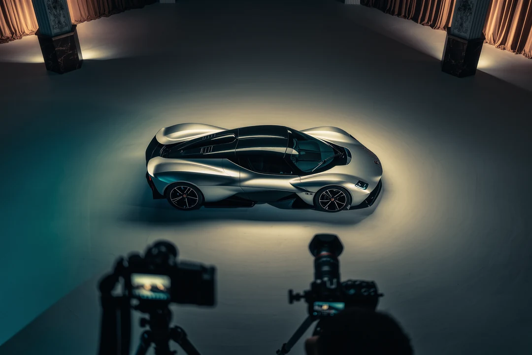 A silver McLaren supercar is parked on a street in front of modern office buildings and trees under a partly cloudy sky.