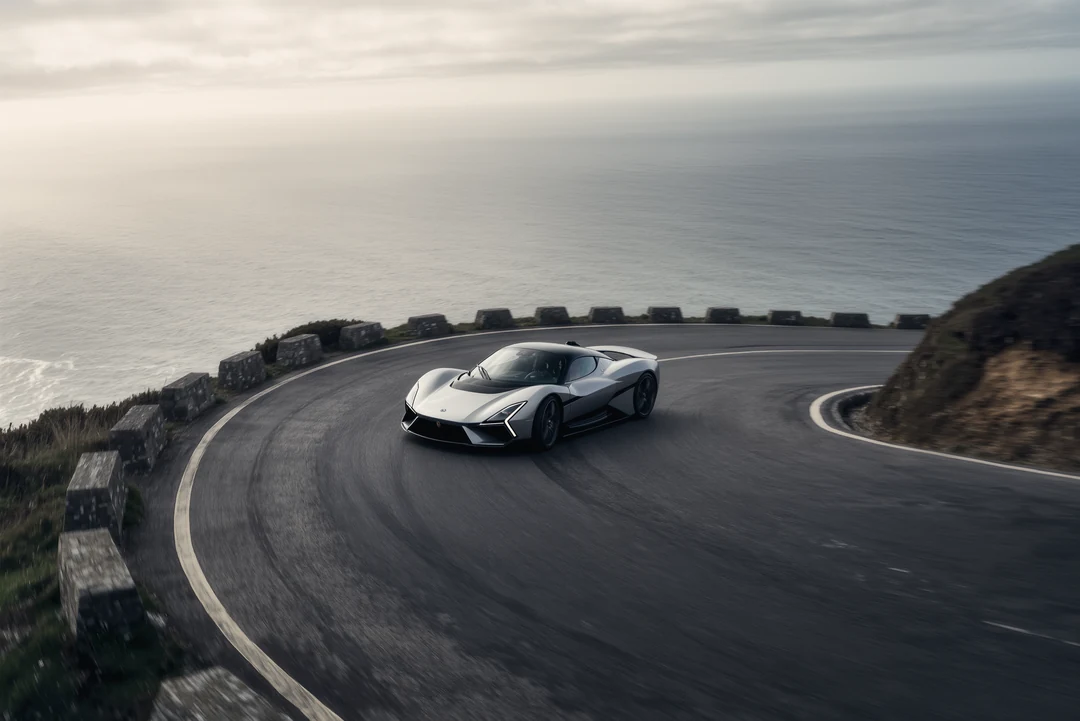 A silver supercar is parked on a paved lot in front of a modern office building, with trees and a fence in the background.