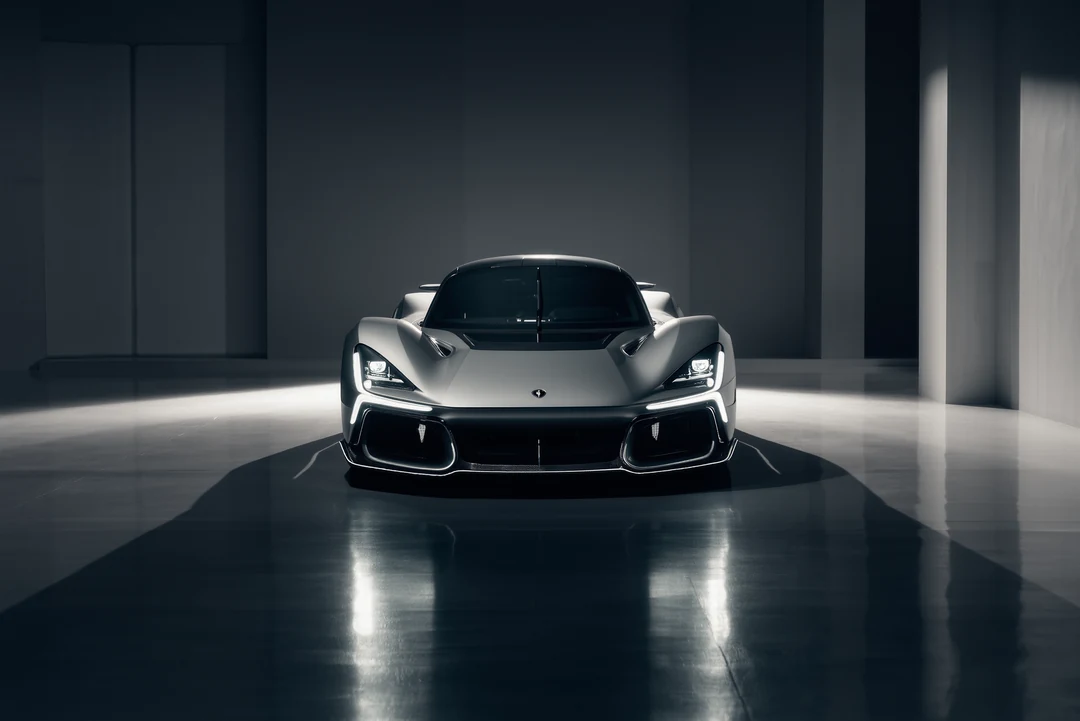 A sleek silver supercar is parked on a street near modern office buildings and trees under a partly cloudy sky.
