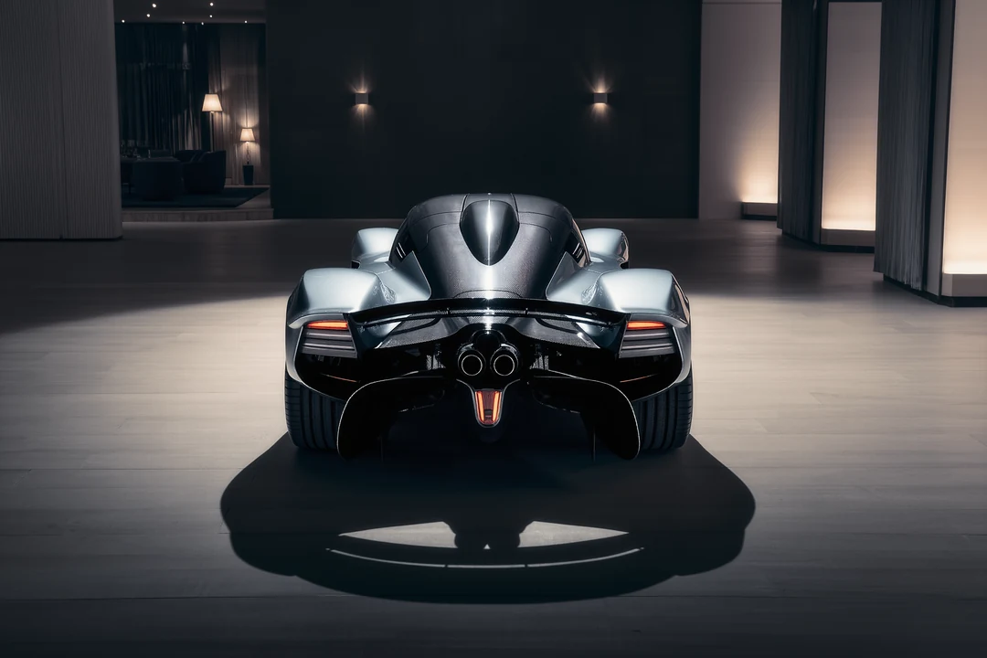 A sleek supercar is parked beside a modern glass building on a racetrack, with sunlight reflecting off the structure.