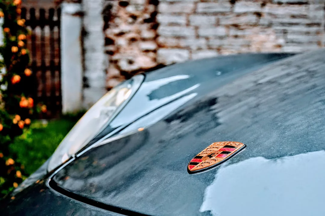 Close-up of a car hood featuring the Porsche logo, one of the most iconic symbols in the world of supercars, with a blurred brick wall and greenery in the background.
